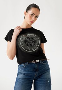 Black t-shirt with a circular design featuring intricate white detailing. Paired with blue jeans and a brown belt with a metallic buckle.