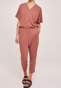 Combinaison rose avec manches courtes, décolleté en V, taille élastique et jambes fuselées. Portée avec des sandales à plateforme noires. Tissu lisse.