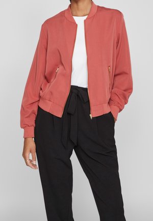 Femme portant une veste bomber corail zippée sur un haut blanc et un pantalon noir taille haute noué avec une ceinture en tissu.
