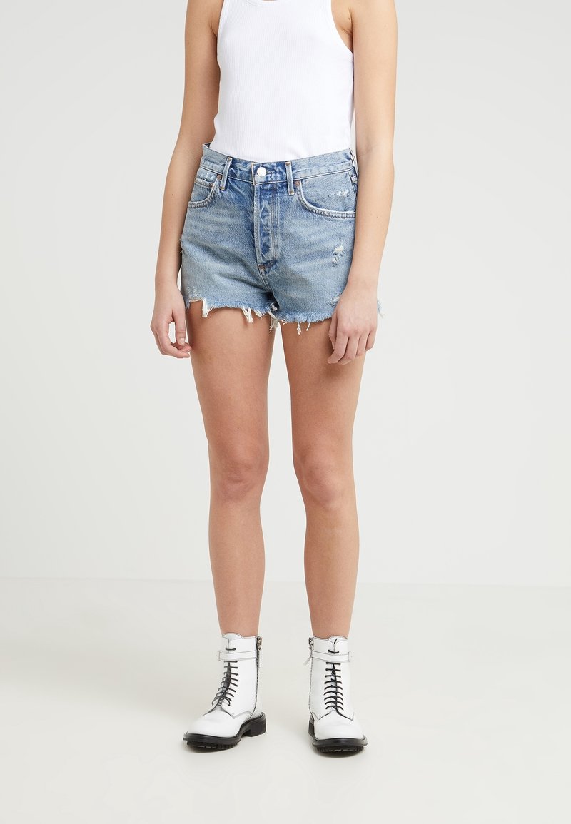 Pantalones cortos de mezclilla de lavado claro con dobladillos deshilachados, diseño de cinco bolsillos y cierre de botón. Combinados con una camiseta sin mangas blanca y botas de encaje negras.