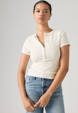 Ung kvinde med mørkt hår iført en hvid kortærmet knapbluse og blå jeans, stående foran en ensfarvet lys baggrund.