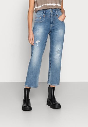 Femme portant un jean court bleu clair usé et des bottines noires, debout avec une main dans la poche devant un fond uni.