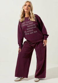 Punainen huppari ja siihen sopivat leveät housut, jotka on valmistettu pehmeästä kankaasta. Hupparissa on painatettuja kaupunkien nimiä vaaleanharmaalla. Vapaa-ajan jalkineet näkyvät.