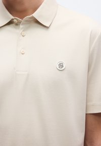 Béžové polo tričko s klasickým golierom, krátkymi rukávmi a bielym kruhovým logom na hrudi. Má zapínanie na dva gombíky. Hladká textúra.