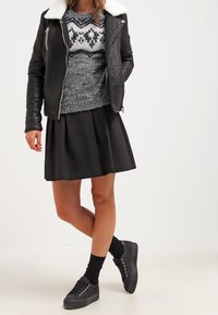 Donna con giacca di pelle nera con colletto bianco, maglione grigio a motivi, gonna plissettata nera, calze nere e sneakers nere, in piedi con le gambe incrociate.