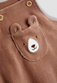 Braune Corduroy-Latzhose mit einer bärenförmigen aufgenähten Tasche, die gestickte Gesichtdetails und Holzknöpfe an den Schultern aufweist.