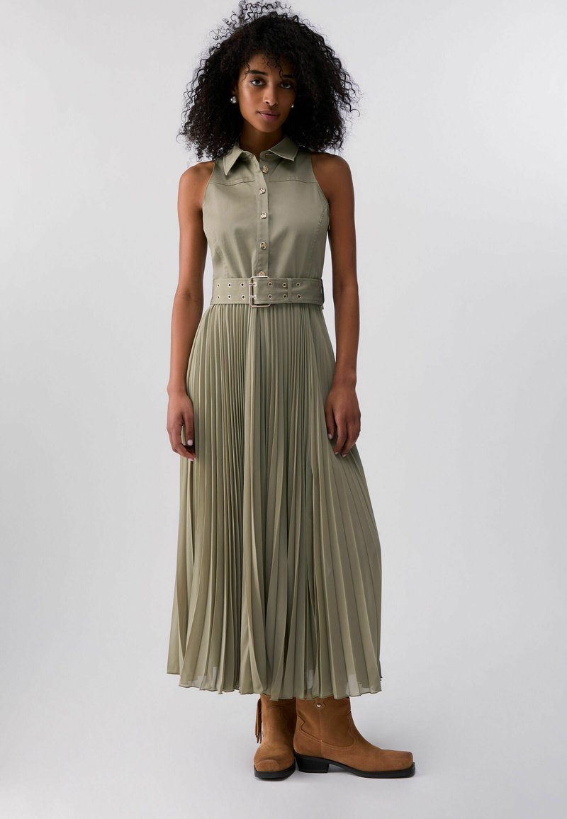 Žena s kučeravými vlasmi v bezrukávových olivovo zelených plisovaných šatách s širokým opaskom a hnedými členkovými topánkami na jednoduchom pozadí.