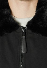 Svart jacka med krage av fuskpäls, med en slät textur, dragkedjestängning och ribbad stickad halsringning.
