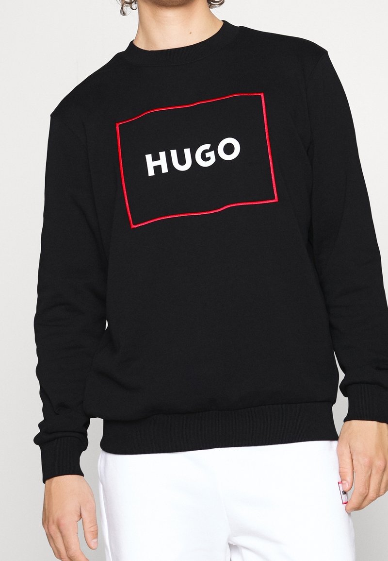 Une personne portant un sweat-shirt noir avec un logo rectangulaire souligné de rouge affichant le mot "HUGO", associé à un pantalon blanc.