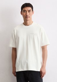 Biała bawełniana koszulka z okrągłym dekoltem, krótkimi rękawami i małym nadrukiem po lewej stronie klatki piersiowej. Noszona z ciemnymi dżinsami na jednolitym tle.