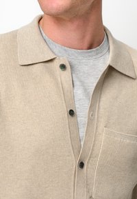 Beige Rippstrick-Cardigan mit einem Kragen, zwei schwarzen Knöpfen und einer Brusttasche, getragen über einem grauen Shirt.