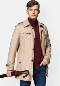 Beżowy płaszcz trench z zapięciem na guziki i naramiennikami, w połączeniu z burgundowym golfem i ciemnoniebieskimi dżinsami. Trzyma czarny parasol.