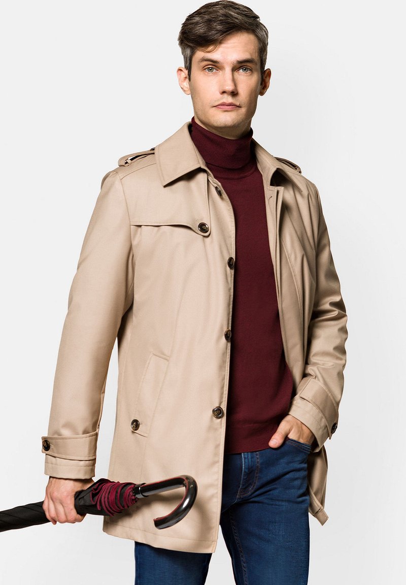 Beżowy płaszcz trench z zapięciem na guziki i naramiennikami, w połączeniu z burgundowym golfem i ciemnoniebieskimi dżinsami. Trzyma czarny parasol.