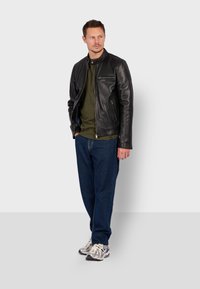 Chaqueta de cuero negra con cuello alzado, cierre delantero, camisa verde debajo, jeans azules y zapatillas grises. Posicionada sobre un fondo claro.