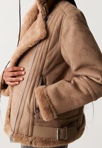 Brown shearling jacket with a soft, textured finish, featuring a wide collar, silver zipper, side pockets, and a belted hem.