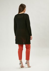 Schwarzes Langarmshirt mit einem Zickzackmuster, kombiniert mit orangefarbenen Hosen und silbernen High Heels, von hinten gesehen.