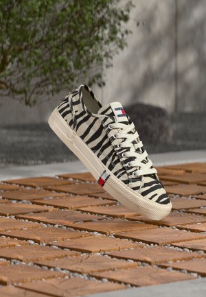 Canvas-sneaker med sort- og hvid zebrastribet mønster, hvide snørebånd og en beige sål, placeret på et trægulv udendørs.