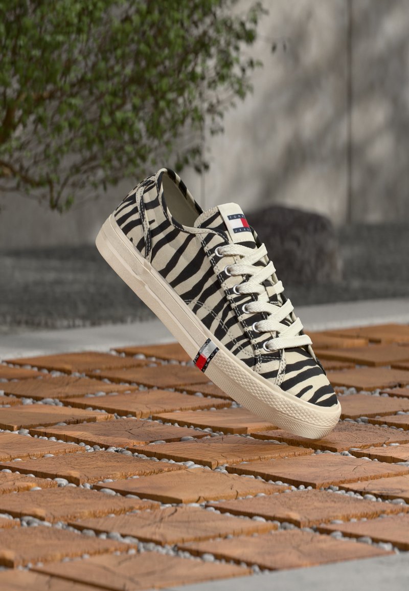 Καμβάς αθλητικού παπουτσιού με ασπρόμαυρες ρίγες ζέβρας, λευκά κορδόνια και μπεζ σόλα, τοποθετημένο πάνω σε ξύλινο δάπεδο εξωτερικού χώρου.