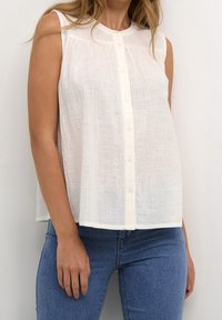 Ærmeløs hvid skjorte med knapper og struktureret stof, båret med blå denimjeans, der viser overkrop og arme mod en ensfarvet baggrund.