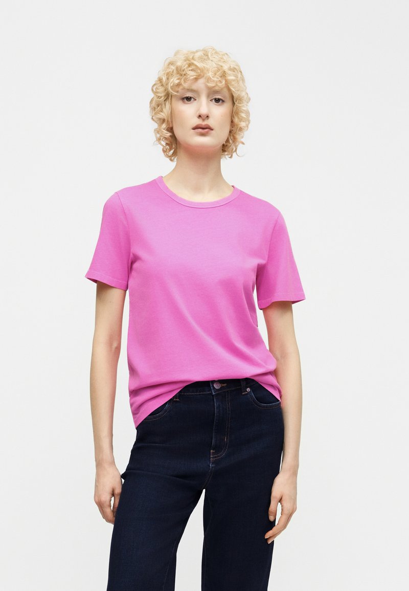 Jeune personne aux cheveux blonds bouclés portant un t-shirt rose vif à manches courtes et un jean bleu foncé sur un fond blanc uni.