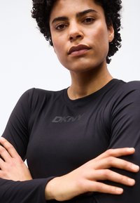 Αυτοπεποίθηση άτομο με κοντά σγουρά μαλλιά φοράει μαύρο μακρυμάνικο πουκάμισο με διακριτικό λογότυπο DKNY, με τα χέρια σταυρωμένα σε λευκό φόντο.