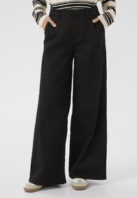 Pantalons noirs à jambes larges en tissu coton, avec des poches latérales, une taille haute et une texture lisse. Portés avec des chaussures claires.