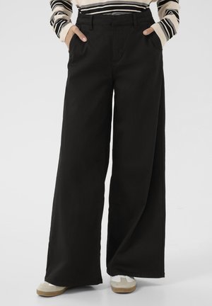 Pantalons noirs à jambes larges en tissu coton, avec des poches latérales, une taille haute et une texture lisse. Portés avec des chaussures claires.
