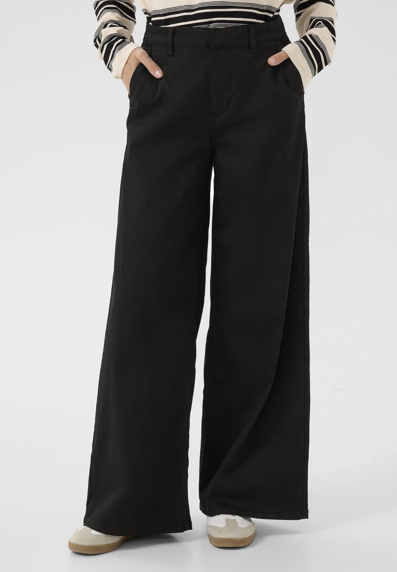 Pantalons noirs à jambes larges en tissu coton, avec des poches latérales, une taille haute et une texture lisse. Portés avec des chaussures claires.