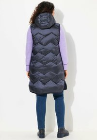 Lång, vadderad marinblå väst med chevronmönster, kviltad design och huva. Bärs över en ljuslila tröja och blå jeans, tillsammans med svarta skor.