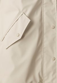 Lys beige yttertøy med en glatt, shiny overflate. Har en skrå lomme og runde trykknapper foran.