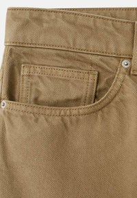 Gros plan sur un tissu en denim beige montrant une poche arrière avec une double couture et des rivets métalliques sur un pantalon.