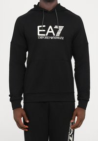 Homme portant un sweat à capuche noir Emporio Armani EA7 et un pantalon de survêtement noir assorti avec texte du logo blanc sur la poitrine et la jambe.