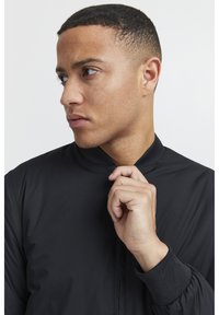 Giacca bomber leggera nera con colletto a bavero, polsini a coste e texture liscia. La mano del modello afferra il colletto.