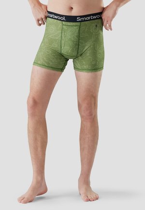 Mężczyzna w zielonych bokserkach w wzory z czarną gumką Smartwool, stojący boso na jednolitym szarym tle.