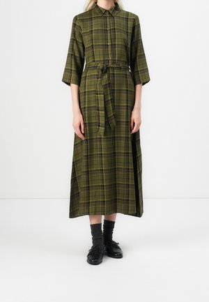 Woman wearing olive green plaid midi dress with buttons, tie waist, three-quarter sleeves, paired with dark socks and black shoes.