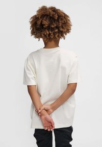 Cremefarvet bomulds T-shirt med korte ærmer, afslappet pasform og rund halsudskæring. Båret af et barn med krøllet hår, hænderne foldet bag ryggen.