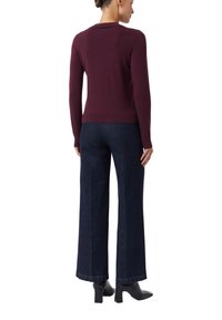 Vrouw in een donkerrode langemouwen trui, donkere blauwe wijde jeans en zwarte haklaarzen, staand met haar rug naar de camera.