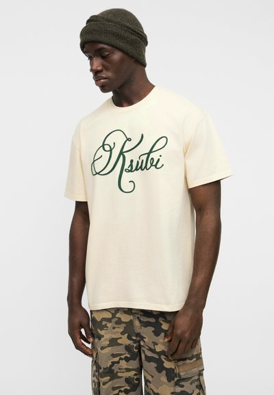 Κρεμ βαμβακερό t-shirt με πράσινο καλλιγραφικό λογότυπο "Ksubi", στρογγυλή λαιμόκοψη και κοντά μανίκια, συνδυασμένο με καμουφλάζ cargo παντελόνια και σκούφο από πλεκτό ύφασμα.