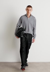 Grauer Reißverschluss-Pullover, dunkle strukturierte Hose, trägt eine schwarze Jacke. Getragen mit schwarzen Schuhen mit roten Akzenten. Einfaches, modernes Design.