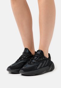 Mustat urheilukengät, joissa on verkkokangas- ja mokkanahkapinta, teksturoitu yksityiskohta, pehmustettu nilkkapanta sekä paksu, kaareva pohjarakenne.