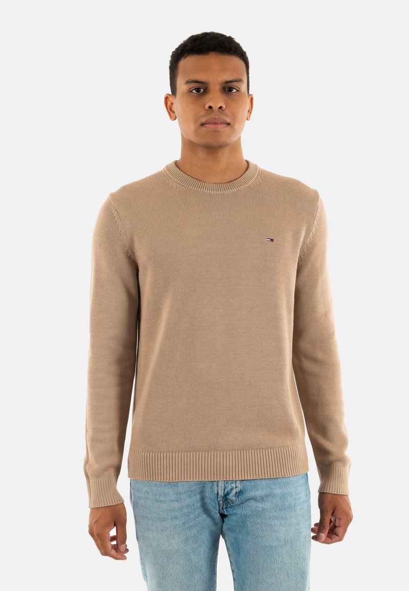 Maglione beige lavorato a maglia con collo a giro, polsini e orlo a coste, con un piccolo logo sul petto, abbinato a jeans azzurri chiari.