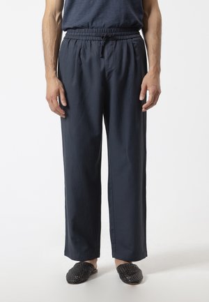 Hombre que lleva pantalones azul marino oscuro con cordón y zapatos negros tejidos sin cordones, de pie frente a un fondo claro y liso.