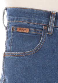 Jeans in denim blu scuro con una finitura testurizzata, caratterizzati da cuciture arancioni e una patch in pelle marrone con il logo sulla vita.