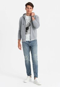 Sweat à capuche zippé gris clair avec une poche frontale, porté sur un t-shirt graphique blanc. Associé à un jean bleu déchiré et des baskets blanches.
