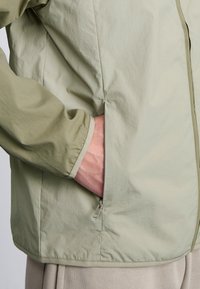 Veste légère vert clair avec une texture lisse et une fermeture éclair ; elle possède une poche latérale et une doublure intérieure contrastante.