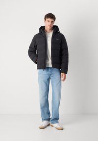 Zwarte pufferjack met ritssluiting en verstelbare capuchon, met een gevoerd ontwerp. Gecombineerd met lichtblauwe jeans en witte sneakers.
