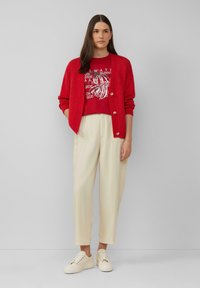 Rode gebreide cardigan over een rood graphic t-shirt, crèmekleurige broek en crèmekleurige sneakers. De outfit heeft knopen op de cardigan en een grafisch tekstontwerp.