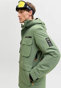 Grüne wasserdichte Jacke mit doppelten Brusttaschen, Seitentaschen und verstellbare Kapuze. Enthält schwarze und orangefarbene Hardware-Akzente.