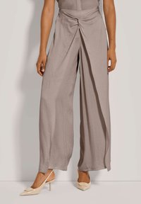 Vrouw in taupekleurige wijde broek met vooroverlapping en beige lage slingback-schoenen, staande tegen een effen achtergrond.
