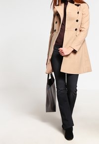 Mujer con abrigo beige, pantalones oscuros y botas, llevando un bolso tote negro, caminando frente a un fondo claro y liso.